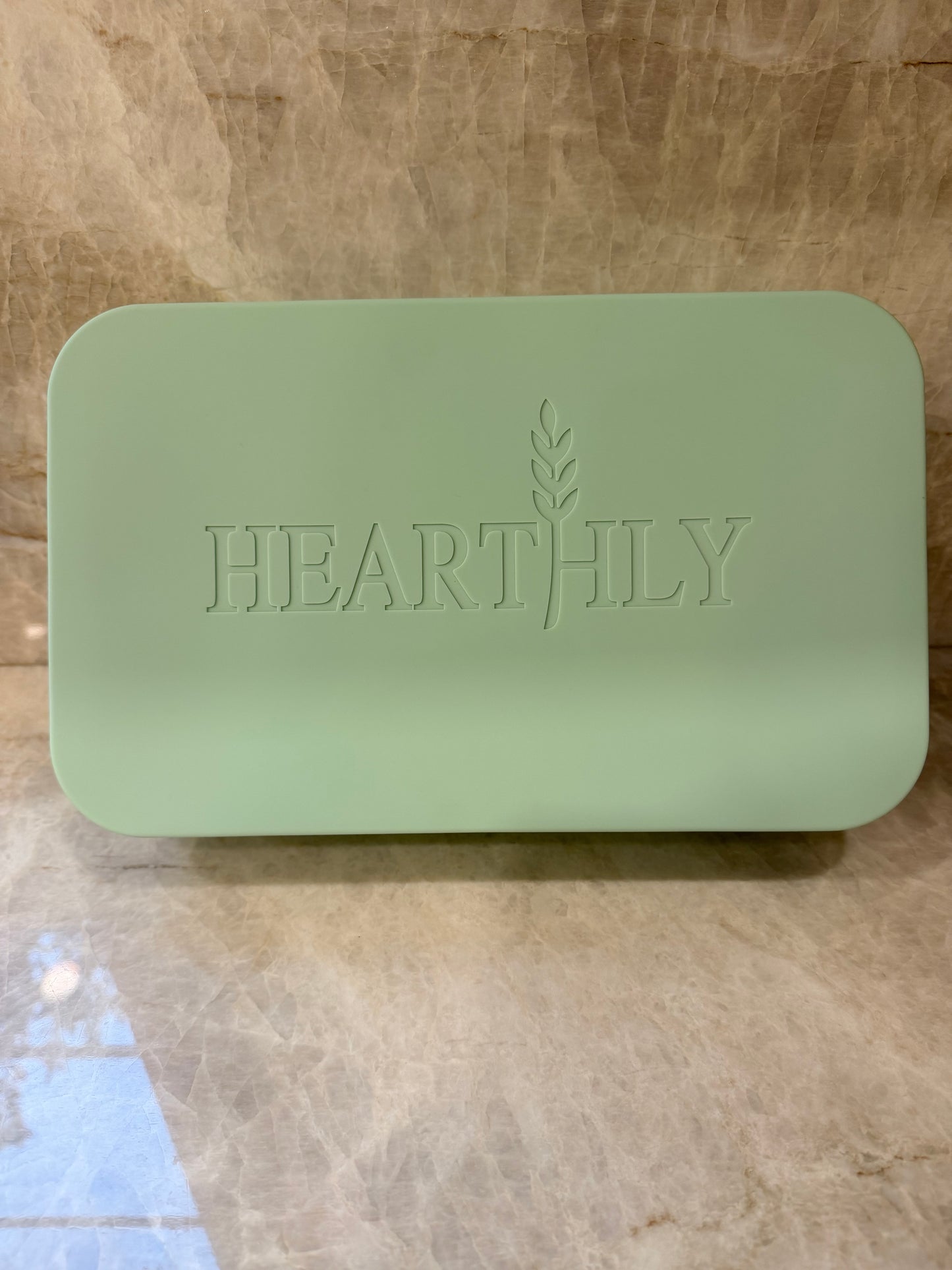 A light green rectangular bar of soap with rounded corners and the brand name 'HEARTHILY' embossed on the front, resting on a beige marble surface.