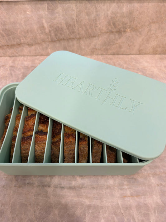 A mint green silicone bread keeper tray with a matching lid embossed with the brand name 'HEARTHILY'. The tray is open, showing several vertical slots holding slices of toasted bread inside.