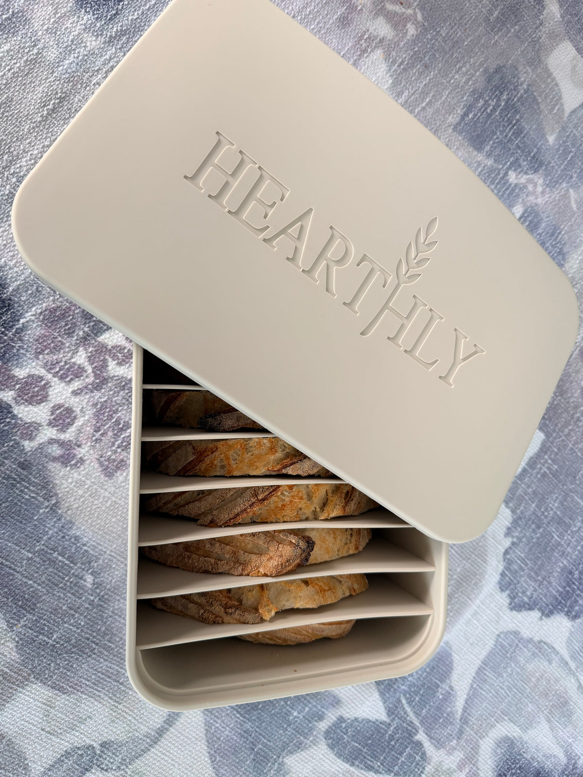 Beige Hearthly bread freezer box with an embossed logo on the lid, holding sliced sourdough bread separated by individual dividers on a blue and white patterned fabric.