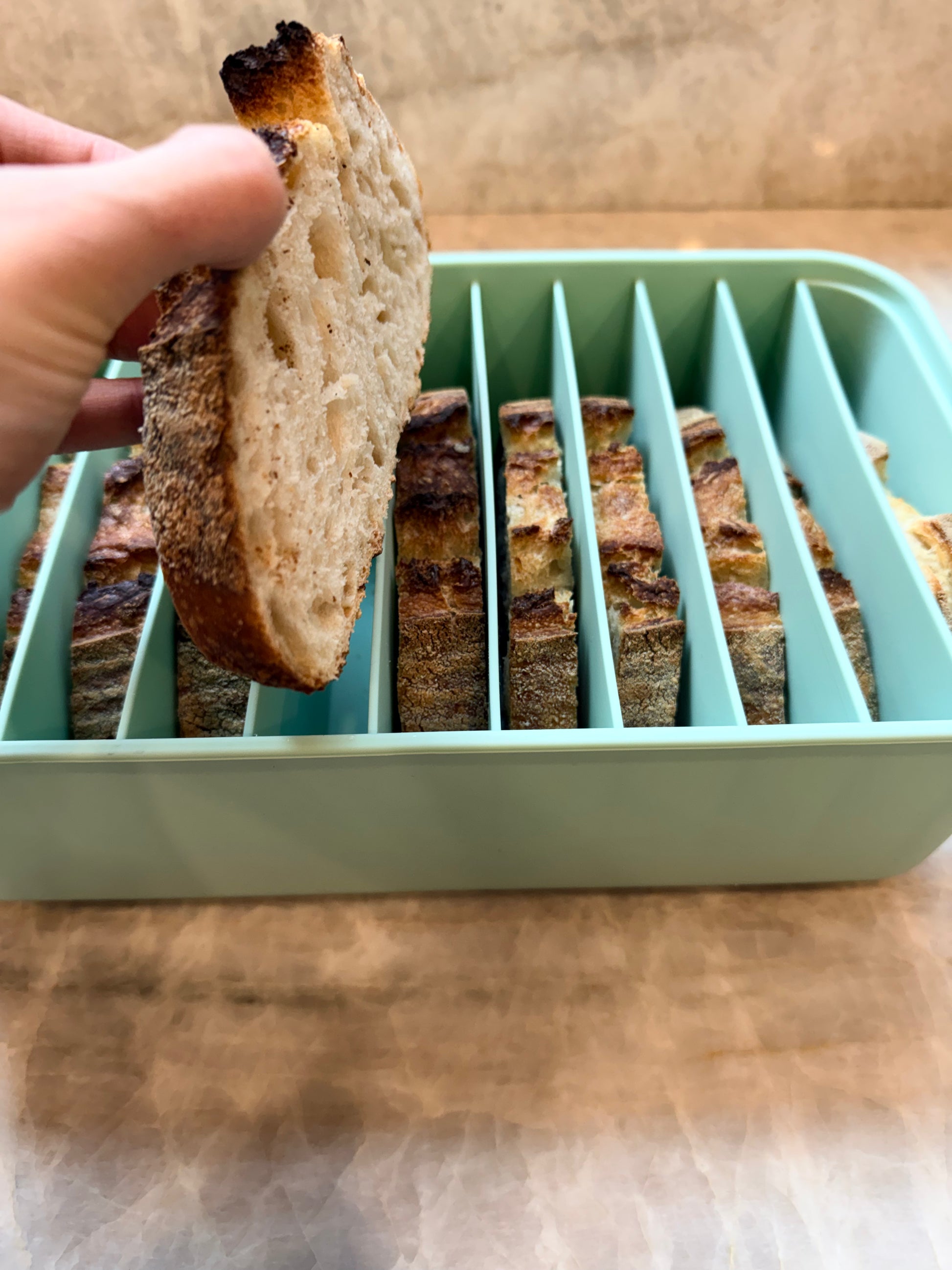 A hand holding a slice of rustic bread over a mint green bread slicer with multiple vertical slots containing other bread slices, set on a wooden surface.