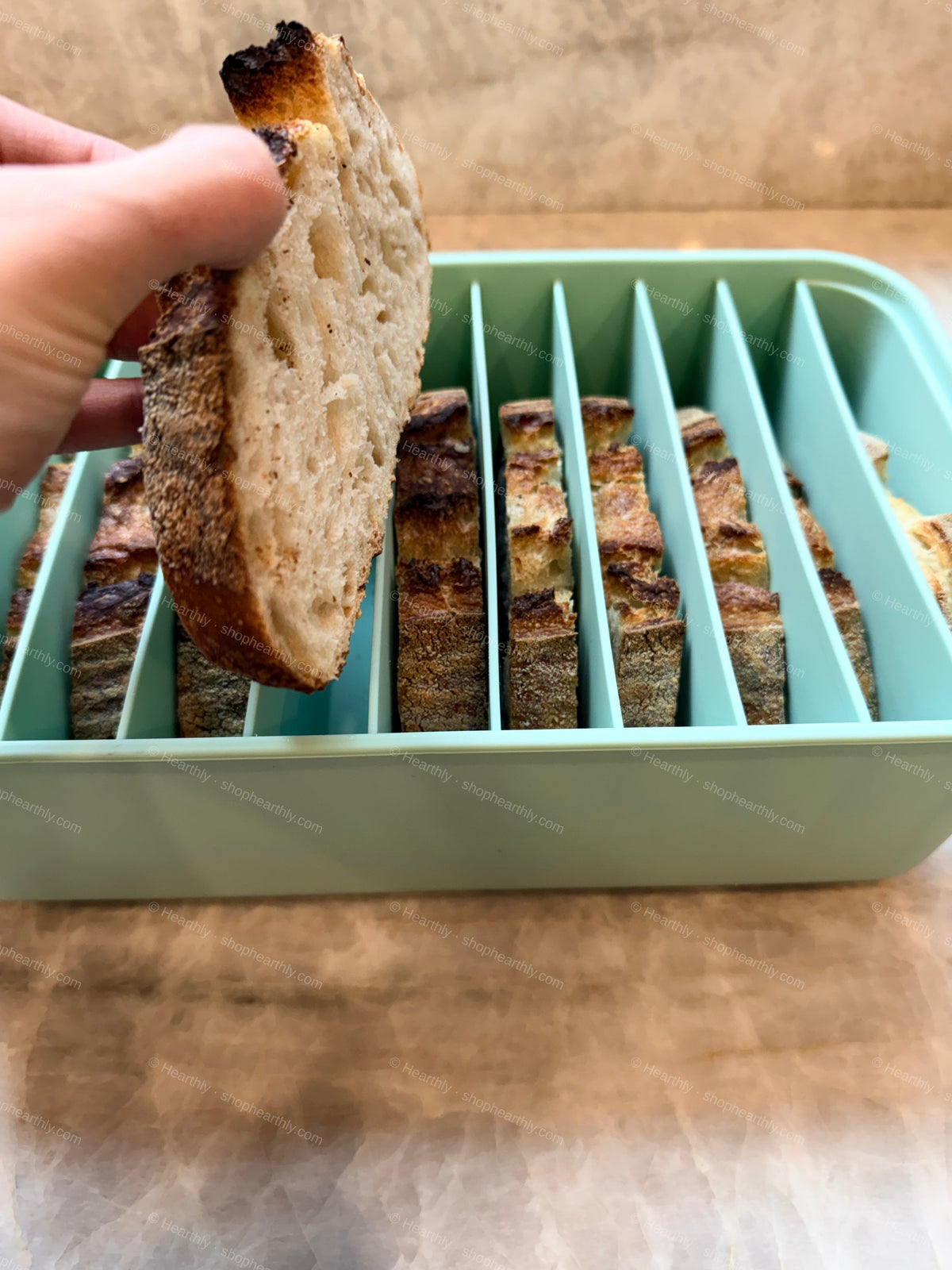 A hand holding a slice of rustic bread over a mint green bread slicer with multiple vertical slots containing other bread slices, set on a wooden surface.